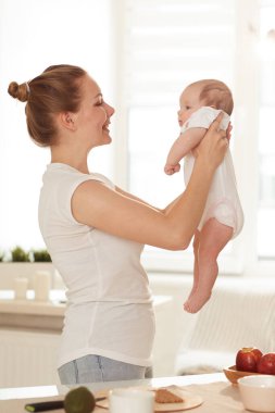 Beyaz tişörtlü, kot pantolonlu, küçük bebeğiyle mutfakta oynayan gülümseyen genç anne.