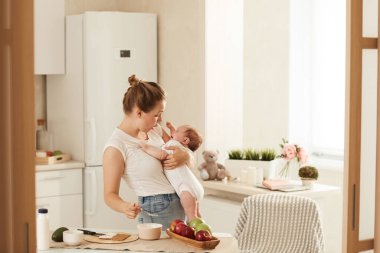 Beyaz tişörtlü genç anne ağlayan ve yemek yemek istemeyen bebeğini neşelendirmeye çalışıyor.