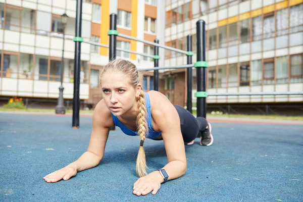 Determined young woman with braid concentrated on elbow plank exercise training alone in yard of apartment house