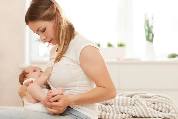 Beautiful young woman sitting on edge of bed and feeding newborn baby with breast milk