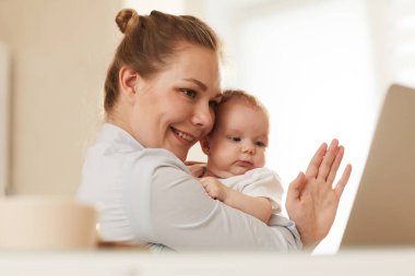 Gülümseyen genç anne bebeğini gösteriyor ve onları Skype üzerinden arayan büyükannesine el sallıyor.