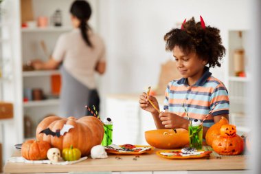 Şeytani boynuzları olan gülümseyen zenci çocuk çeşitli Cadılar Bayramı şekerleriyle tezgahta duruyor ve marmelat solucanı yiyor.