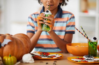 Gecelik tişörtlü zenci çocuğun yemekle tezgahta dikilmesi ve oyuncak gözle Cadılar Bayramı kokteyli içmesi.