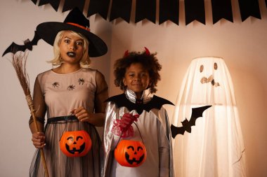 Kafası karışmış cadı şapkalı anne portresi ve Cadılar Bayramı partisi için süslenmiş şeytan kostümü giymiş yakışıklı Afro-Amerikan oğlu.