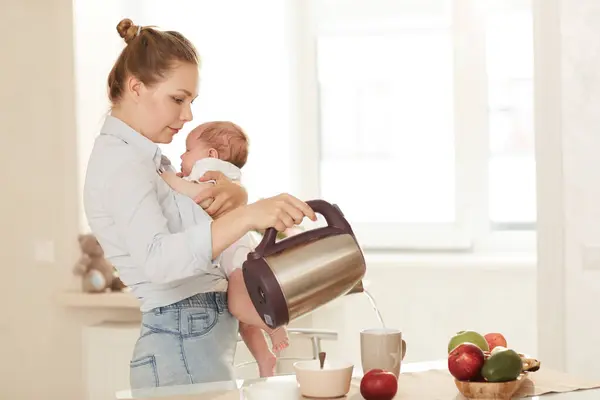 Young mother in blue shirt and jeans standing in kitchen with baby in her arms and making tea