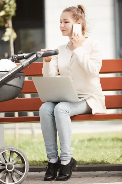 Young mother in white coat working outside on her smartphone and laptop while baby sleeping in stroller
