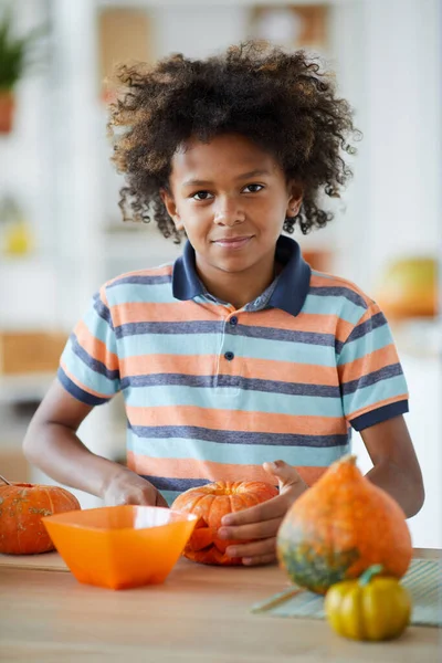 Portrait of content black boy with Afro hairstyle wearing stripped tshirt standing at counter and carving pumpkin