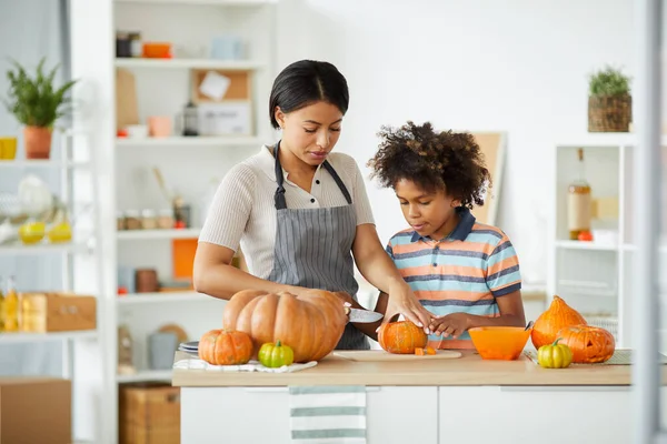 Young beautiful black mother in apron using knife while teaching son to carve Halloween pumpkin
