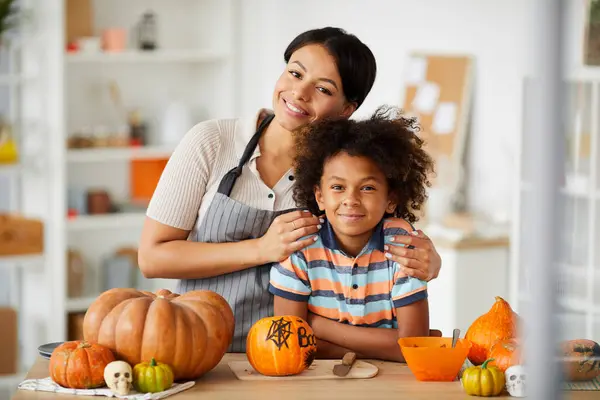 Smiling loving young black mother in apron embracing son while posing with him in Halloween workshop