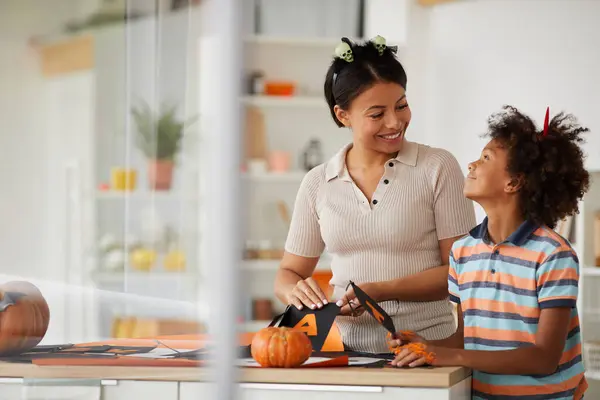 Cheerful young black mother and cute son with Afro hairstyle standing at counter and preparing paper decorations for Halloween with pleasure