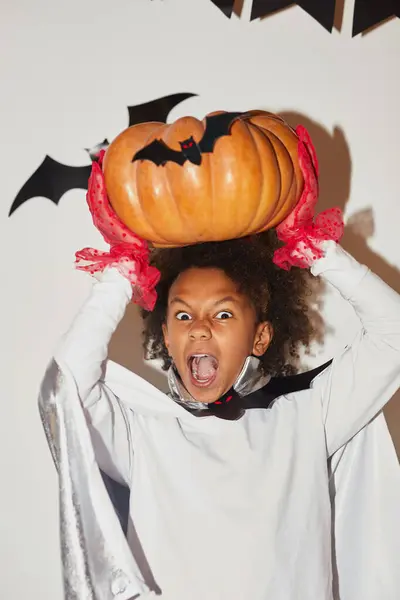 Angry emotional African boy in cape and red gloves holding large pumpkin above head and screaming at camera