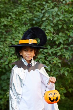 Sihirbaz kıyafetli gülümseyen siyah çocuk parkta duruyor ve kameraya bakarken şeker sepeti tutuyor.