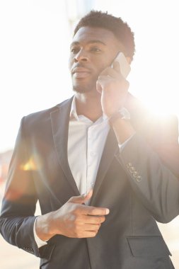 Kendine güvenen takım elbiseli genç bir iş adamı güneş ışığında duruyor ve cep telefonuyla projeyi tartışıyor.