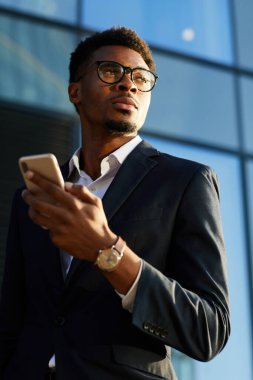 Ciddi iç gözlemci stil sahibi takım elbiseli siyahi iş adamı dışarıda duruyor ve akıllı telefondan internette sörf yapıyor.