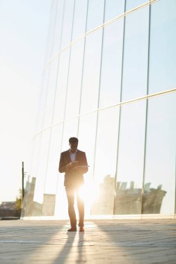 Genç Afro-Amerikalı iş adamı cam binaya karşı duruyor ve akıllı telefonu hareket halinde kullanırken dizüstü bilgisayarını tutuyor.