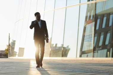 Takım elbiseli çağdaş siyahi yönetici finans bölgesinde yürüyor ve müşteriyle telefonla konuşuyor.