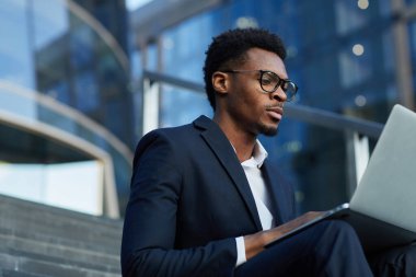 Gözlüklü ciddi siyahi yönetici ofis binasına karşı merdivenlerde oturup dizüstü bilgisayar kullanarak iş sunumuna odaklanmış durumda.