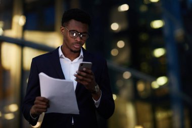 Gerçekten meşgul, gözlüklü genç bir adam gece ofis binasına karşı duruyor ve elinde kağıtlarla internetten bilgi topluyor.