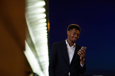 Pozitif yakışıklı takım elbiseli genç siyahi iş adamı. Karanlık şehir caddesinde ilerliyor ve telefonla mesajları kontrol ediyor.