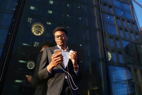 Below view of pensive young black businessman in wired earphones standing by modern building and using mobile mail on phone