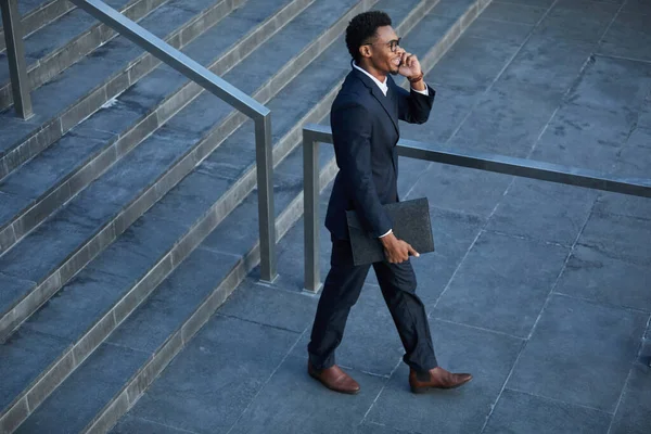 Positive young black lawyer in stylish suit moving down stairs and talking by phone while leaving court