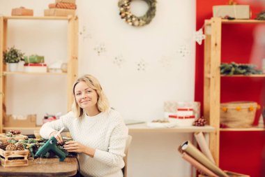 Beyaz kazaklı, gülümseyen sarışın bir kadının portresi atölyede oturuyor ve Noel çelengi için doğal malzemelerle çalışıyor.