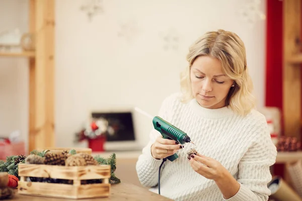 Concentrated young woman with blond hair using glue gun while applying hot glue on cone to attach it to Christmas wreath in craft studio