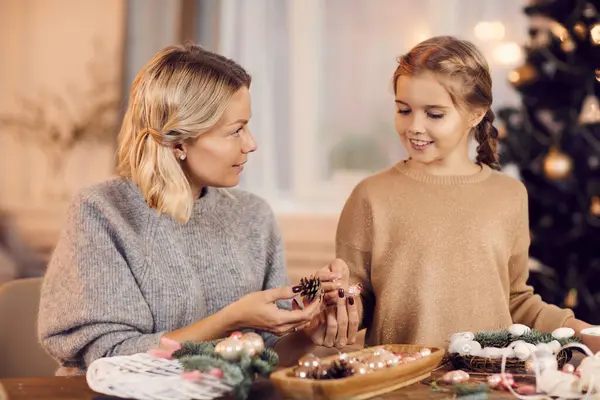 Attractive young blonde mother sitting at table and discussing natural decorations for Christmas wreath with daughter at home