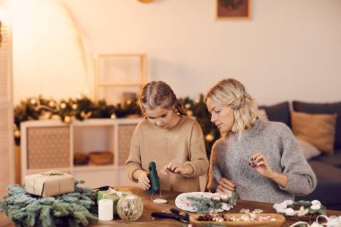 Gri kazaklı genç anne oturma odasındaki tahta masada oturuyor ve kızına tatil çelengi yapmayı öğretiyor.