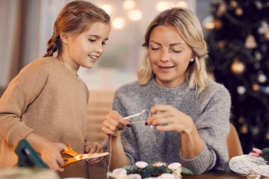 Oturma odasında Noel çelengi üzerinde çalışan genç ve mutlu bir aile. Anne kurdeleyi ölçüyor, kızı kesiyor.