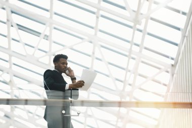 Takım elbiseli ciddi dalgın bir Afrikalı yönetici müşteriyle telefonda konuşurken tırabzanlara yaslanıyor ve evraklara bakıyor.