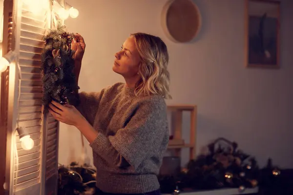 Content young woman inspired with Christmas enjoying decorating home: she hanging beautiful wreath on folding screen