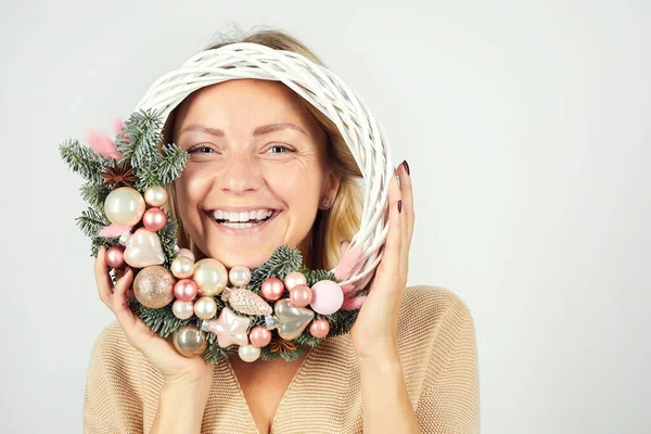 Portrait of happy playful young woman standing against white background and looking through artificial Christmas wreath