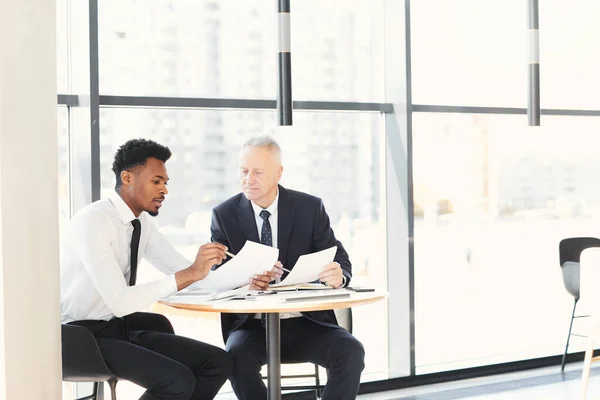 Young African-American forecast expert sitting at table in business cafe and explaining report data to mature colleague