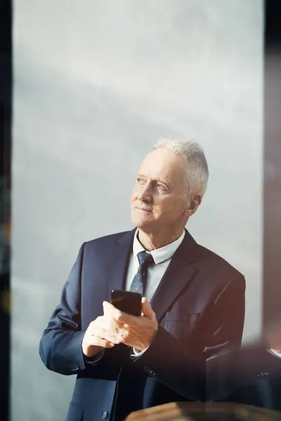 Content dreamy gray-haired businessman in suit standing in lobby and using gadget app for planning time