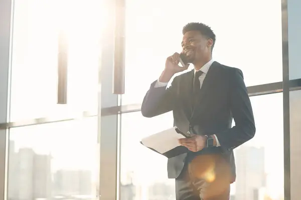Content confident Afro-American manager in suit standing against window and answering clients questions by phone