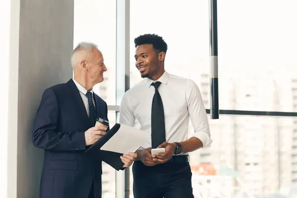 Positive confident senior businessman with coffee cup holding clipboard with papers and discussing project implementation with young black PR manager