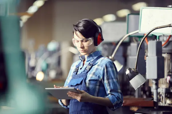 Serious busy young female worker in ear protectors and safety goggles using digital tablet while making notes on processes of production shop