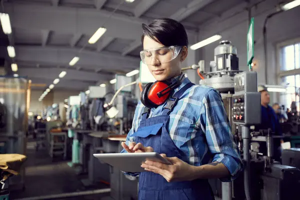 Busy young factory woman in blue checkered shirt and overall standing in industrial shop with machines and adding information into tablet