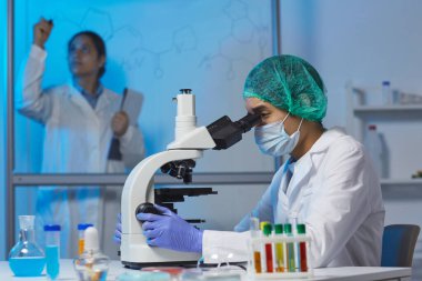 Cerrahi şapkalı ve beyaz şapkalı konsantre genç bilim adamı masada oturuyor ve laboratuvarda kimyasal araştırmalar yaparken mikroskop kullanıyor.