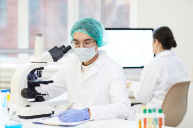 Ciddi bir Asyalı genç laboratuvar araştırmacısı, steril iş kıyafetleri bölümünde mikroskopla oturuyor ve örnek hakkında kayıt tutuyor.