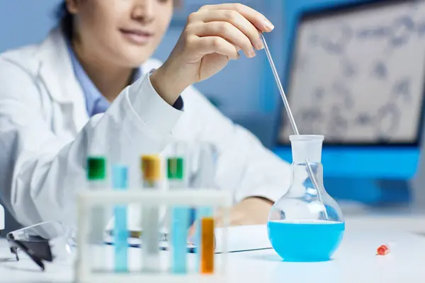 Close-up of content female lab worker in white coat sitting at table and using pipette and beaker while holding experiment in chemical laboratory