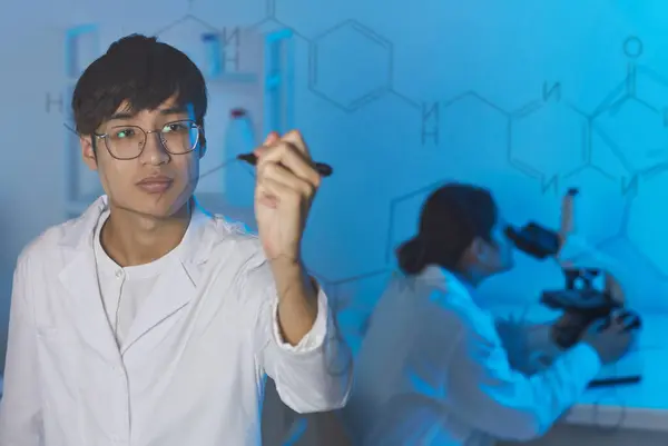 Behind glassy board view of serious young Asian chemist in lab coat writing down chemical formula on glass board
