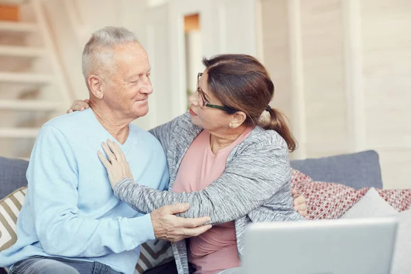 Concerned loving senior wife sitting on sofa and embracing her husband while asking him about health