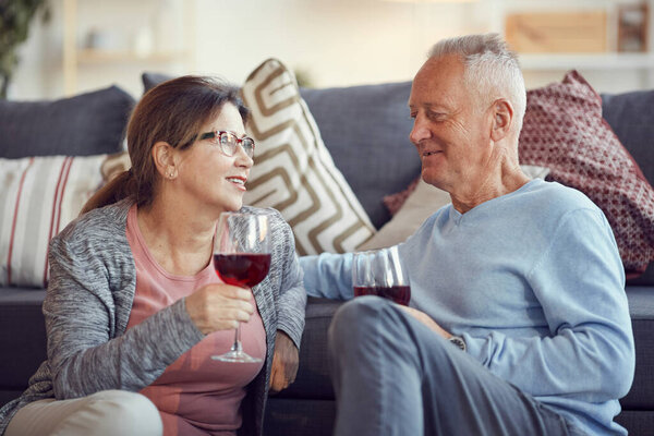 Happy handsome gray-haired man in sweater leaning on sofa and drinking wine while having romantic night with elderly wife