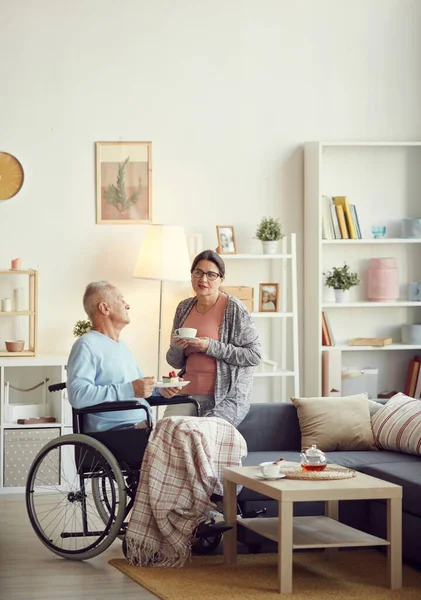 Serious mature woman in eyeglasses sitting on sofa in living room and holding cup of tea while talking to husband in wheelchair