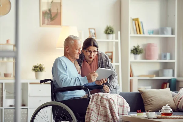 Senior man with legs covered with blanket sitting in wheelchair and showing new mobile app on tablet to surprised wife