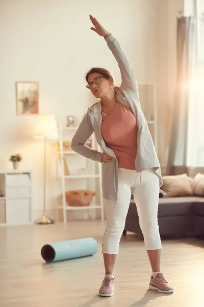 Serious purposeful senior woman in leggings standing in living room and doing side bend exercise alone
