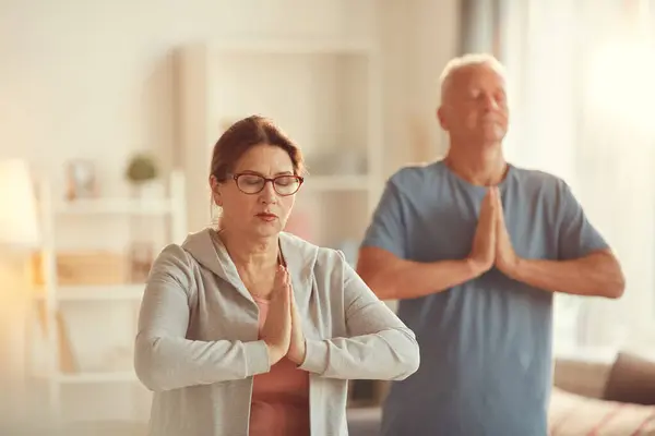 Serene aged couple concentrated on meditation standing in living room and making Namaste gesture