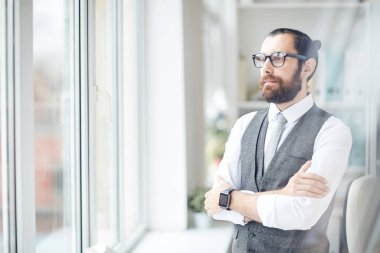 Ciddi, kasıtlı, sakallı, gri yelekli iş adamı modern ofiste dikilip pencereden dışarı bakıyor.